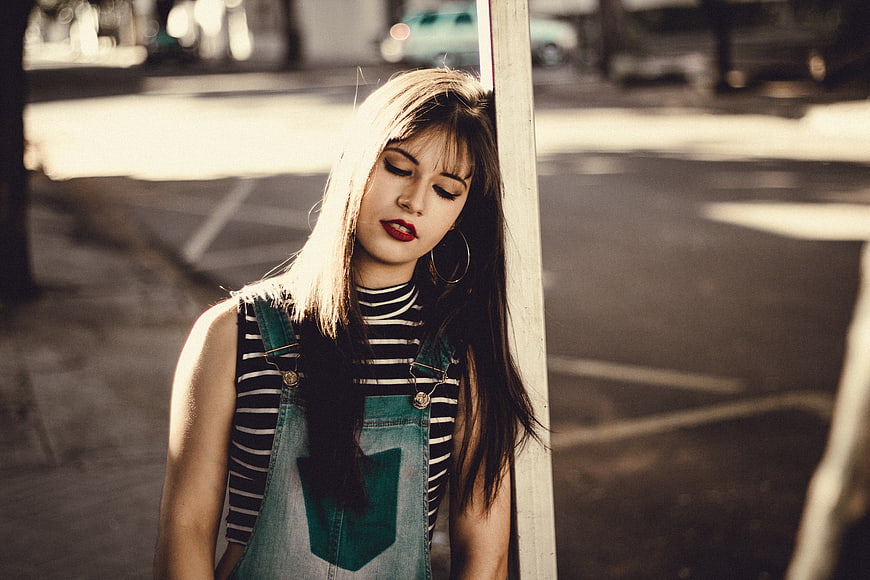 Young woman in denim overalls leaning against a post in sunlight