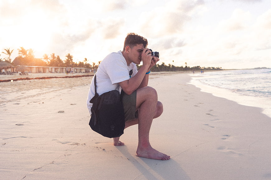 Young Photographer Squatting on Beach at Sunset