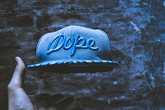 Hand tossing blue snapback hat with Dope text against brick wall