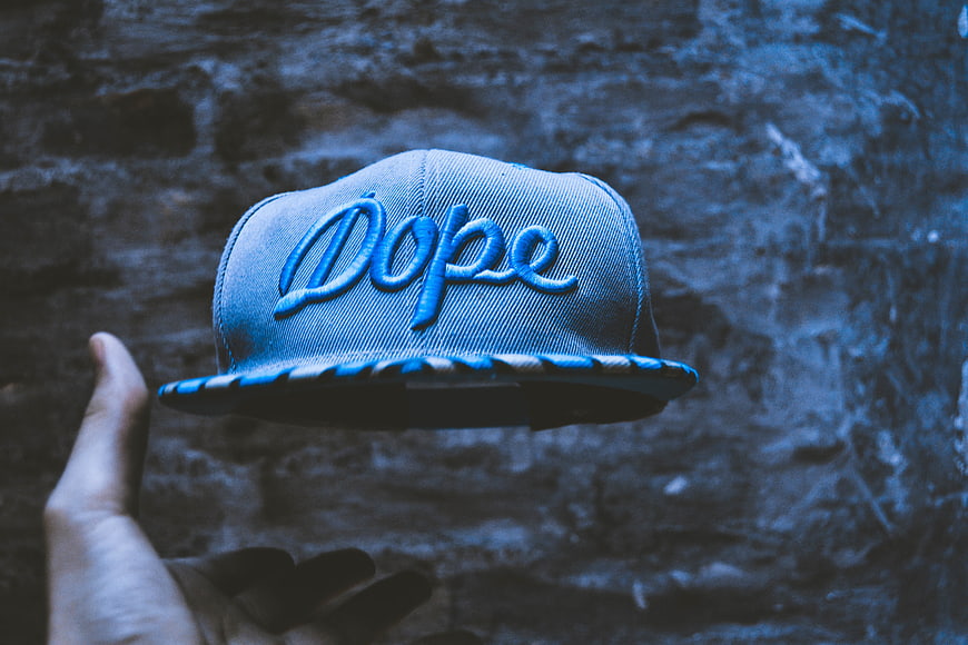 Hand tossing blue snapback hat with Dope text against brick wall