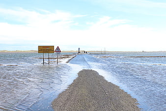 Flooded gravel causeway with danger warning signs in coastal landscape