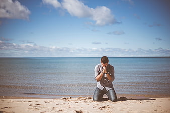 Man kneeling and praying on sandy beach by ocean