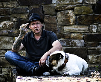 Tattooed man in hat sitting with bulldog outdoors