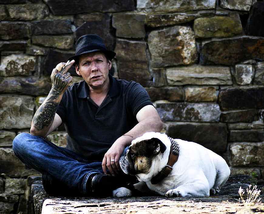 Tattooed man in hat sitting with bulldog outdoors