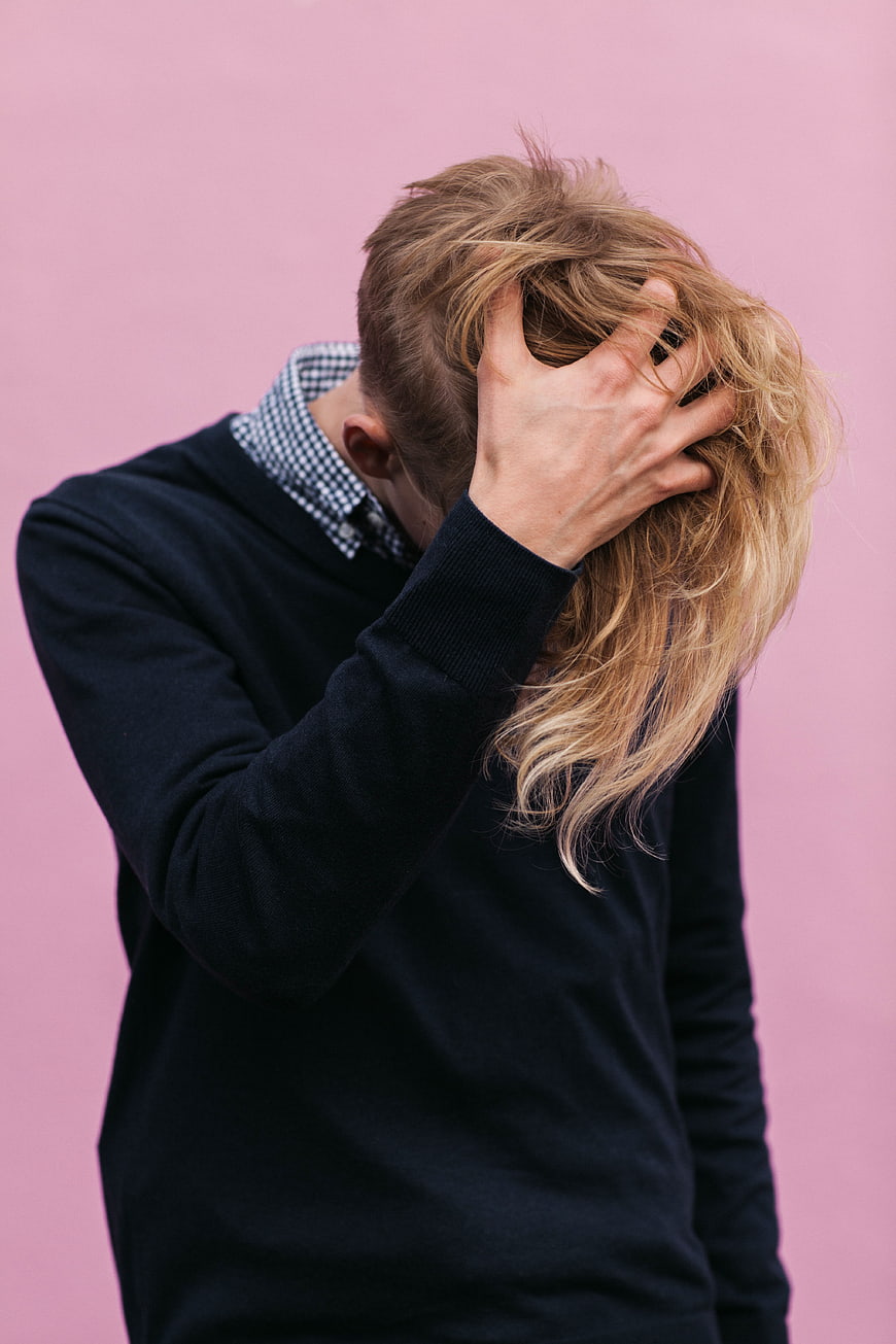 Man with blonde mullet hiding face in hand against pink background