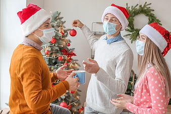 Friends decorating Christmas tree wearing face masks and Santa hats