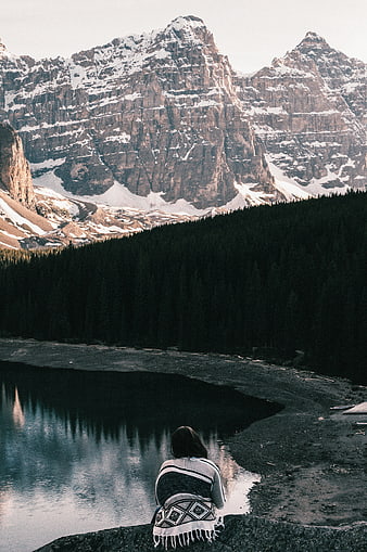 Woman wrapped in blanket sitting by mountain lake