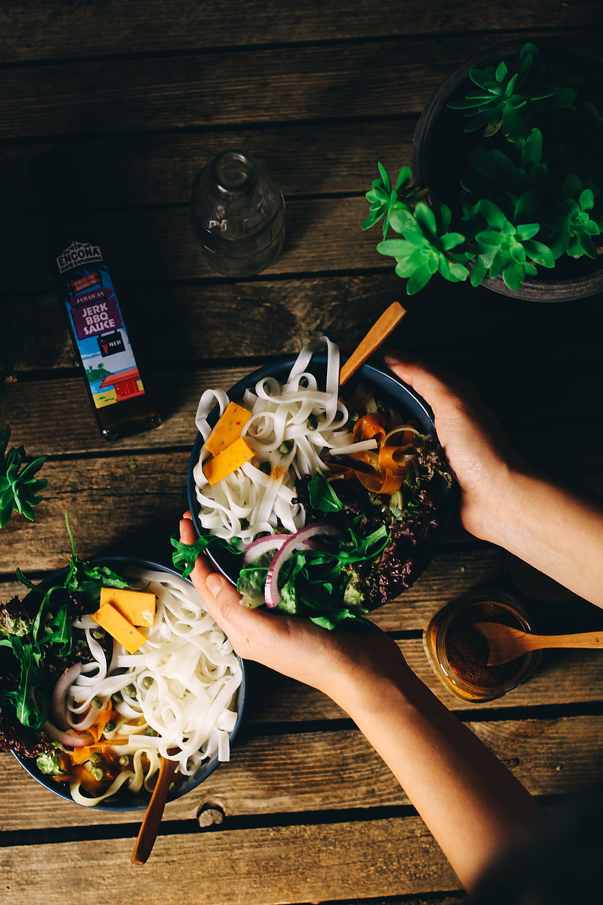 Top view of hands holding vegan noodle bowls on rustic wood