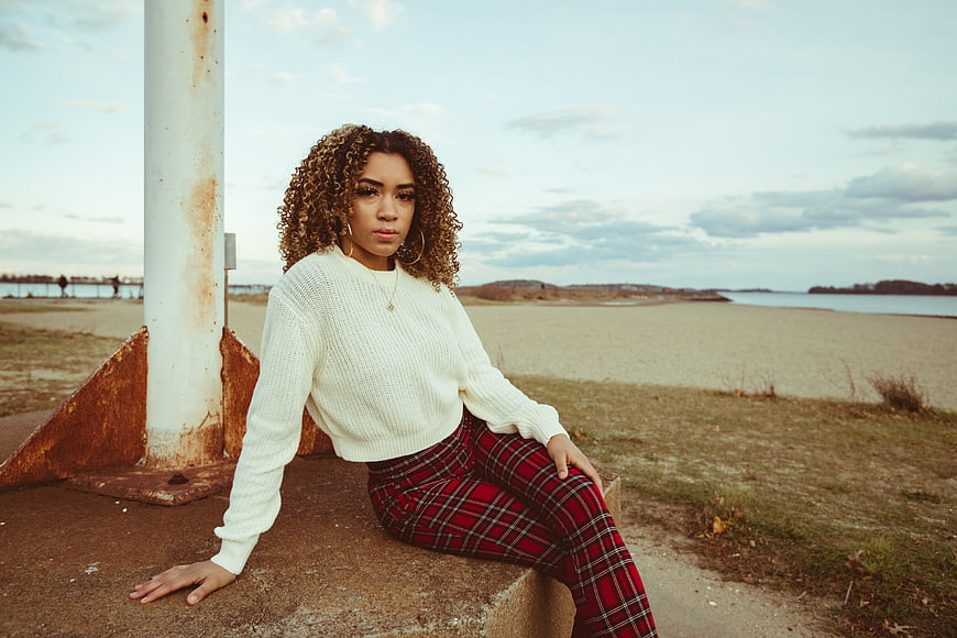 Woman in White Sweater and Plaid Pants Sitting by the Beach
