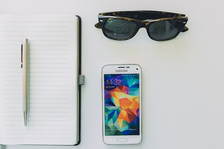 Flat lay workspace with notebook, pen, sunglasses, and smartphone on white background