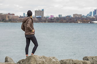 Woman in Brown Puffer Jacket Standing on Rocks by Water