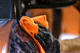 Gloved hand wiping water droplets from a black car with an orange microfiber cloth