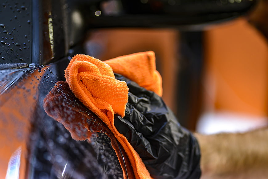 Gloved hand wiping water droplets from a black car with an orange microfiber cloth