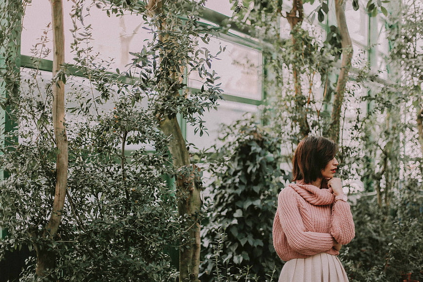 Woman in Pink Sweater Standing in Lush Greenhouse Garden