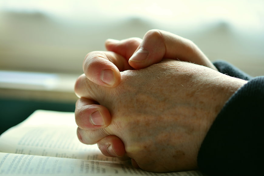 Close-up of elderly hands clasped in prayer over an open book