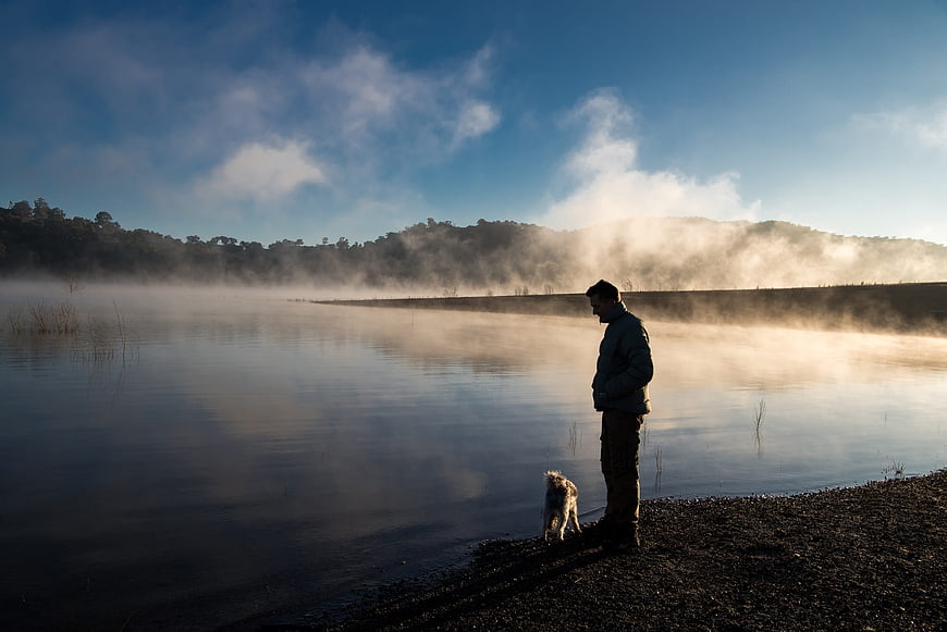 Man and Dog Standing by Misty Lake at Sunrise