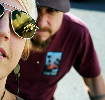 Close-up selfie of woman with sunglasses and man in background