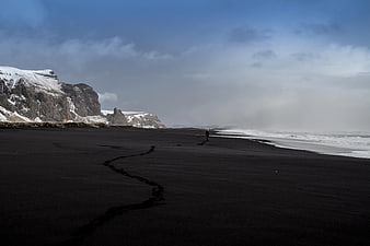 Lone Figure Walking on Black Sand Beach Near Snowy Cliffs