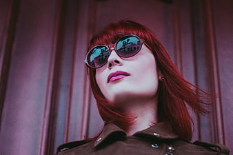 Stylish woman with red hair and sunglasses against purple wall