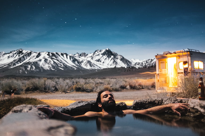 Man relaxing in hot spring with mountains and camper at night