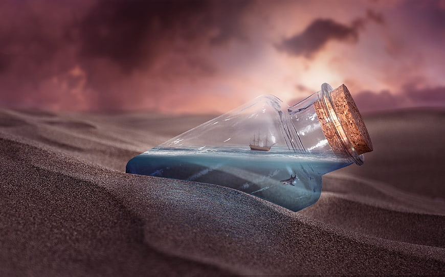 Glass bottle with miniature ship on desert sand at sunset