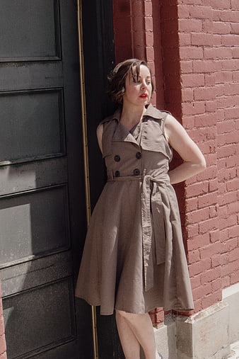 Woman in beige trench dress leaning against red brick wall