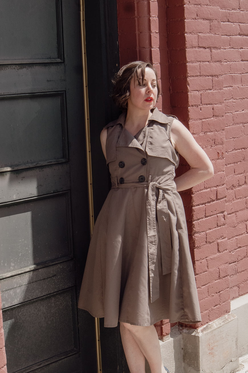 Woman in beige trench dress leaning against red brick wall
