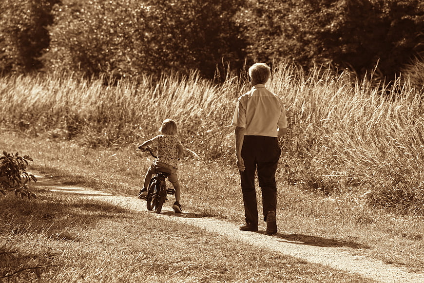 Senior man walking behind child riding bicycle on rural dirt path