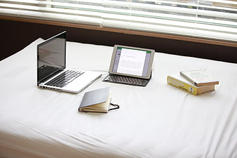 Laptop and tablet workspace on white bed with books