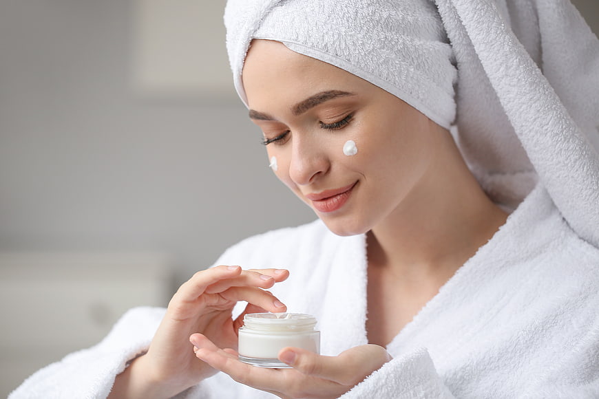 Woman applying face cream in white bathrobe and towel