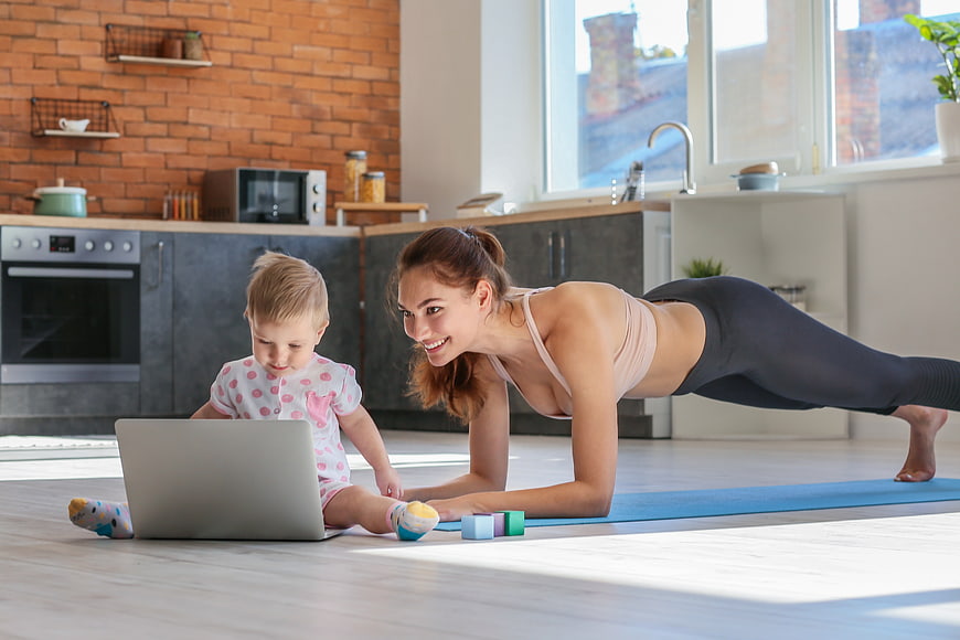 Mother doing yoga plank while toddler watches laptop