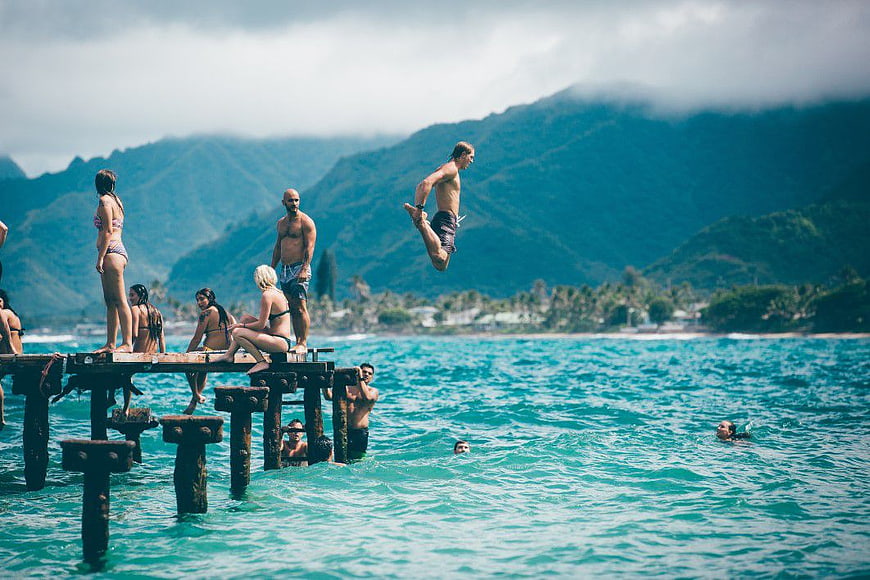Man jumping off wooden pier into turquoise ocean with mountains