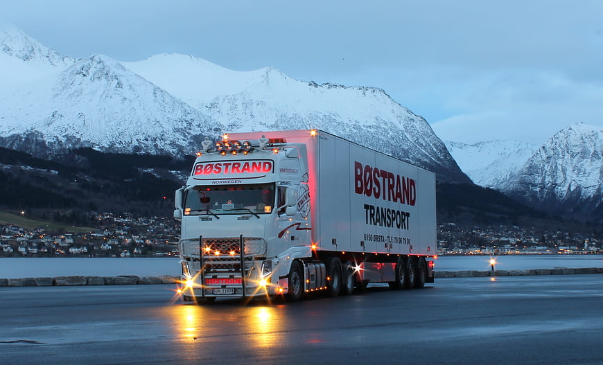 White Volvo Transport Truck Driving on Wet Road with Snowy Mountains