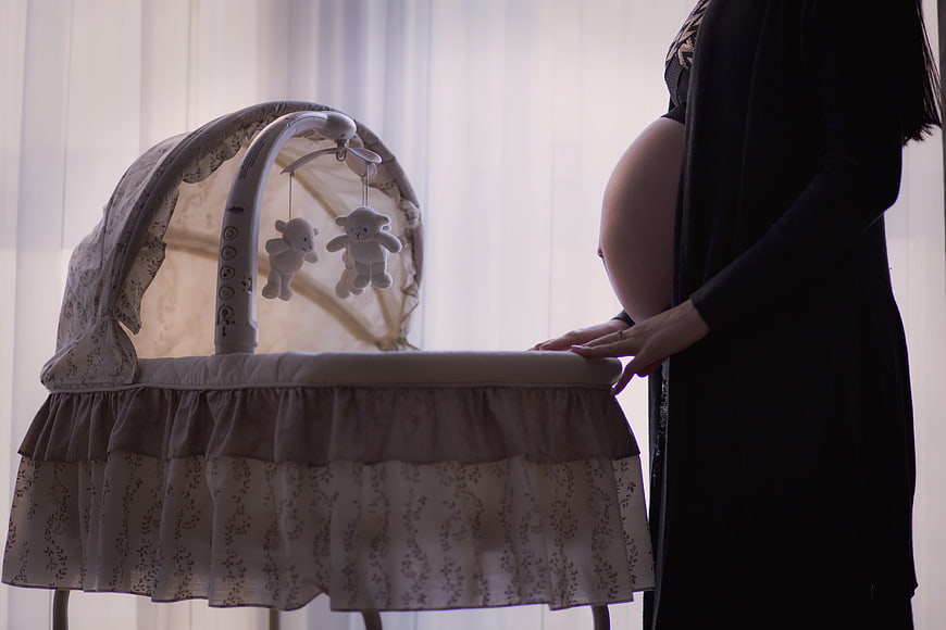 Pregnant woman standing beside a baby bassinet with mobile