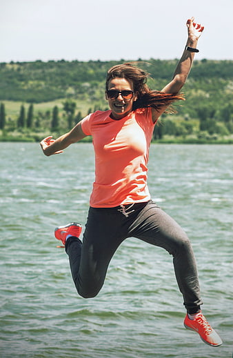 Joyful Woman Jumping In Air By Lake Wearing Orange Sportswear