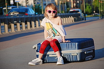 Little girl with pink sunglasses sitting on blue suitcase on city street