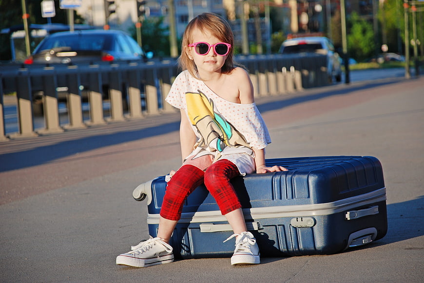 Little girl with pink sunglasses sitting on blue suitcase on city street