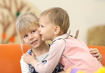Toddler whispering secret to smiling blonde girl on orange sofa