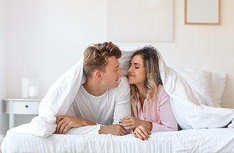 Happy young couple smiling under white duvet in bright bedroom