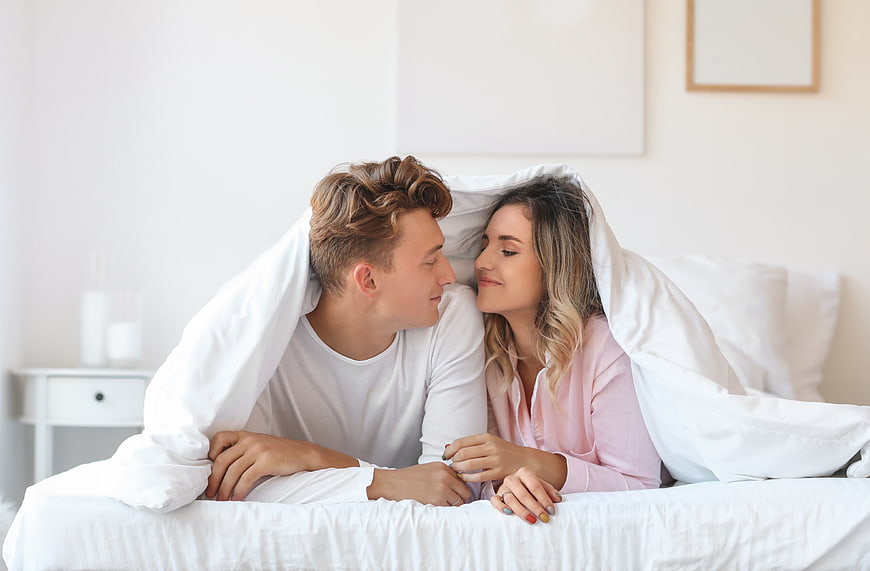 Happy young couple smiling under white duvet in bright bedroom