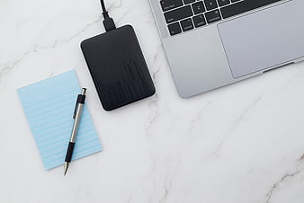 Flat lay of laptop, external hard drive, and notepad on marble
