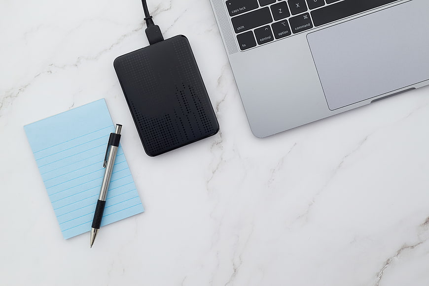 Flat lay of laptop, external hard drive, and notepad on marble