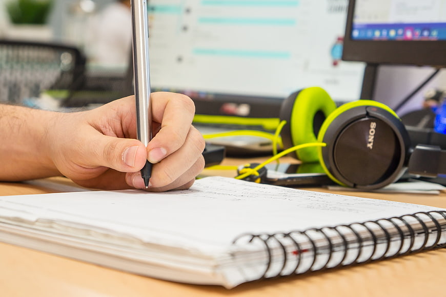 Man Writing Notes In Spiral Notebook At Office Desk