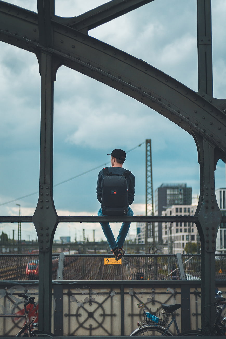 Man with backpack sitting on bridge railing overlooking city