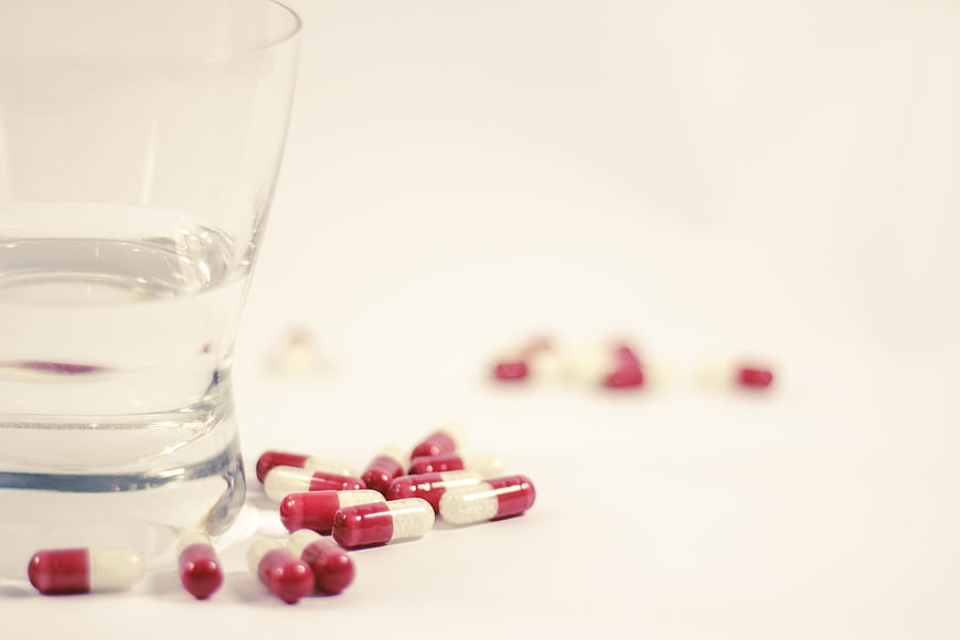 Red and white capsules scattered next to a glass of water on a white background