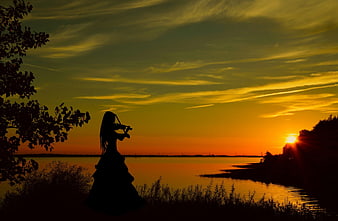 Silhouette of woman playing violin at sunset by river