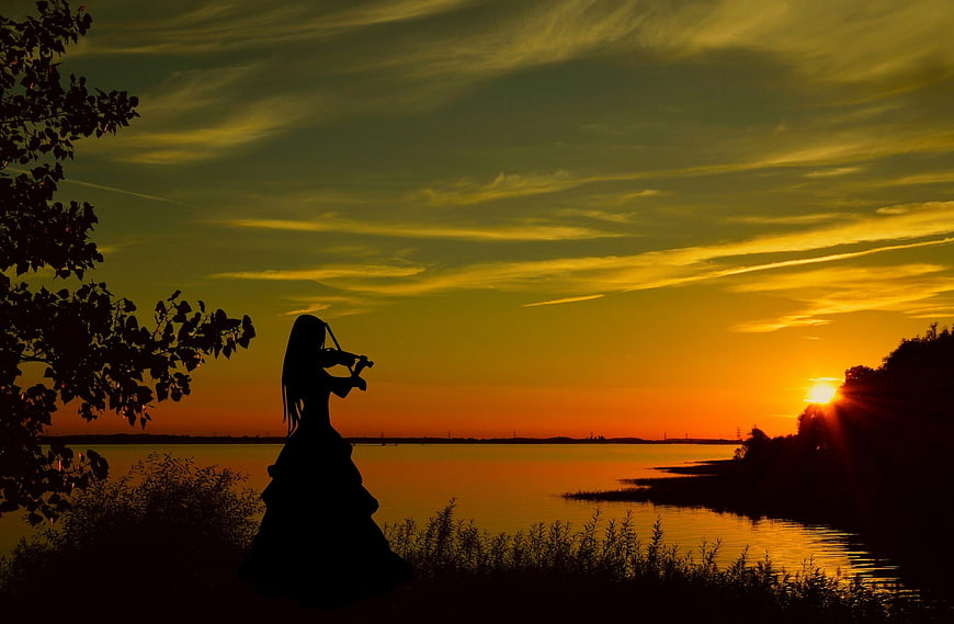 Silhouette of woman playing violin at sunset by river