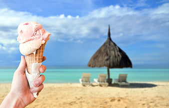 Hand holding ice cream cone on tropical beach with thatched umbrella