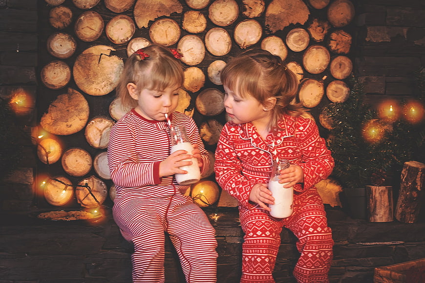 Two young girls in red pajamas drinking milk by a Christmas tree
