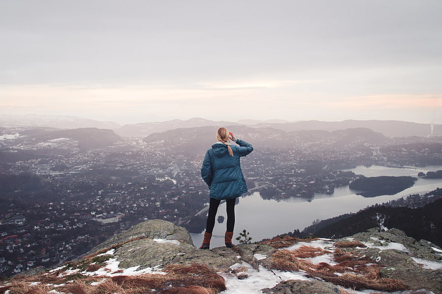 Woman in Blue Jacket Standing on Snowy Mountain Peak Overlooking City